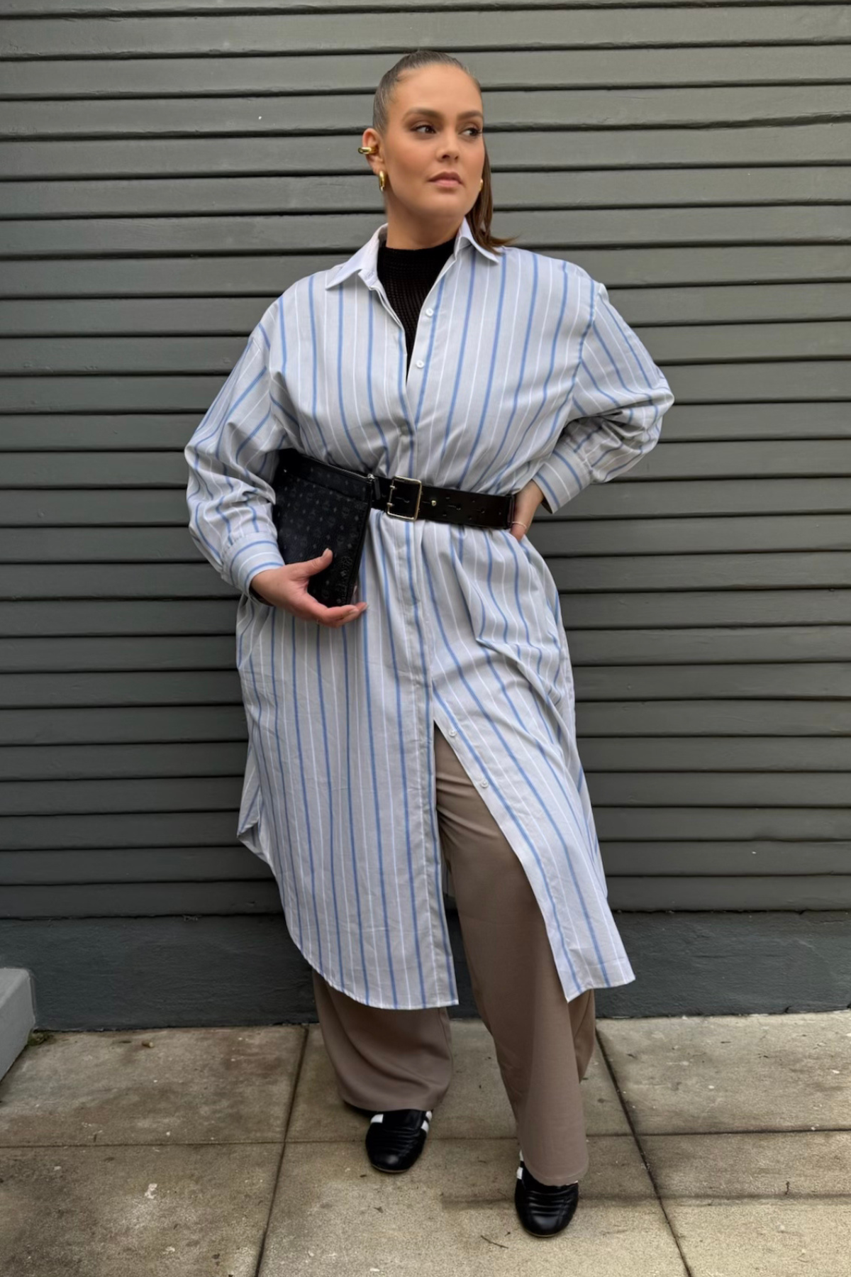 Person_wearing_a_long_striped_shirt_over_a_dress_with_a_belt_standing_against_a_gray_wall..png